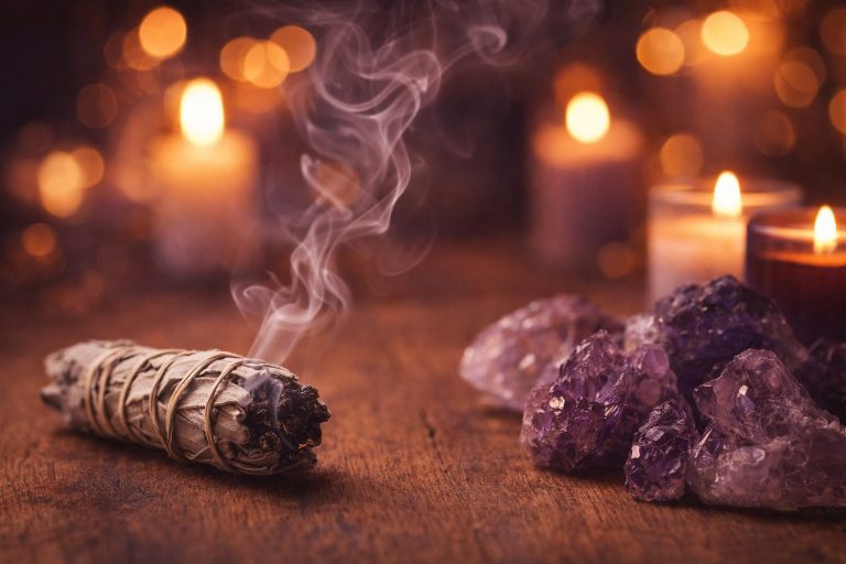 Sauge blanche fumante à côté de cristaux d’améthyste sur une surface en bois, éclairée par des bougies aux lumières violettes et ambrées, ambiance spirituelle et apaisante.