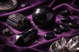 Cristaux noirs élégants — sphère d’obsidienne, tourmaline brute et onyx poli — disposés sur un tissu violet profond, éclairage doux avec ombres marquées, ambiance mystique et luxueuse.