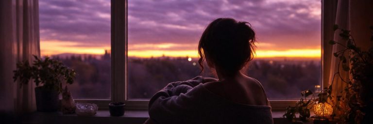 Femme vue de dos assise face à une fenêtre, regardant un ciel crépusculaire violet et doré, ambiance calme, mélancolique et apaisante dans une lumière intérieure tamisée.