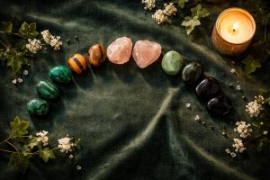 Vue de dessus d’un flat lay luxueux sur velours vert forêt : arc de pierres naturelles (malachite, œil de tigre, quartz rose, aventurine, obsidienne), bougie dorée allumée à droite, petites fleurs blanches et feuilles de lierre, lumière dorée douce et ambiance élégante.