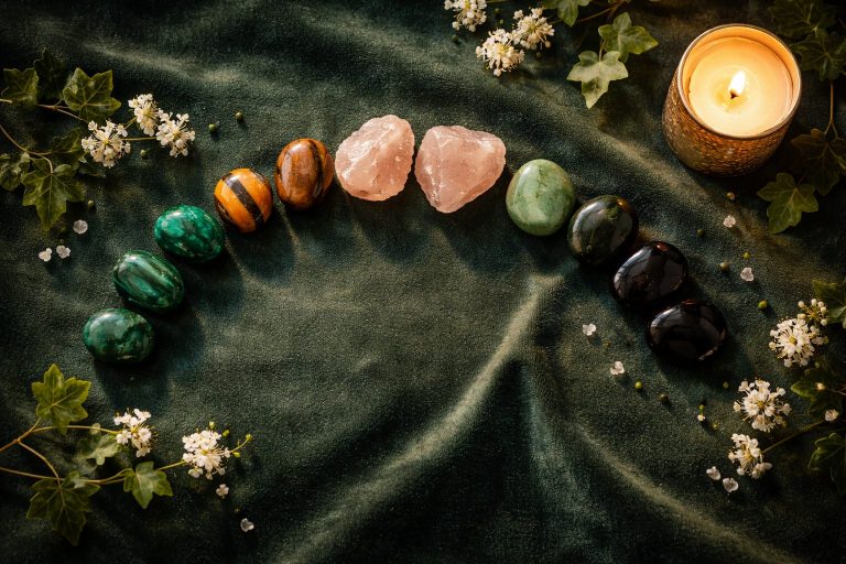 Vue de dessus d’un flat lay luxueux sur velours vert forêt : arc de pierres naturelles (malachite, œil de tigre, quartz rose, aventurine, obsidienne), bougie dorée allumée à droite, petites fleurs blanches et feuilles de lierre, lumière dorée douce et ambiance élégante.